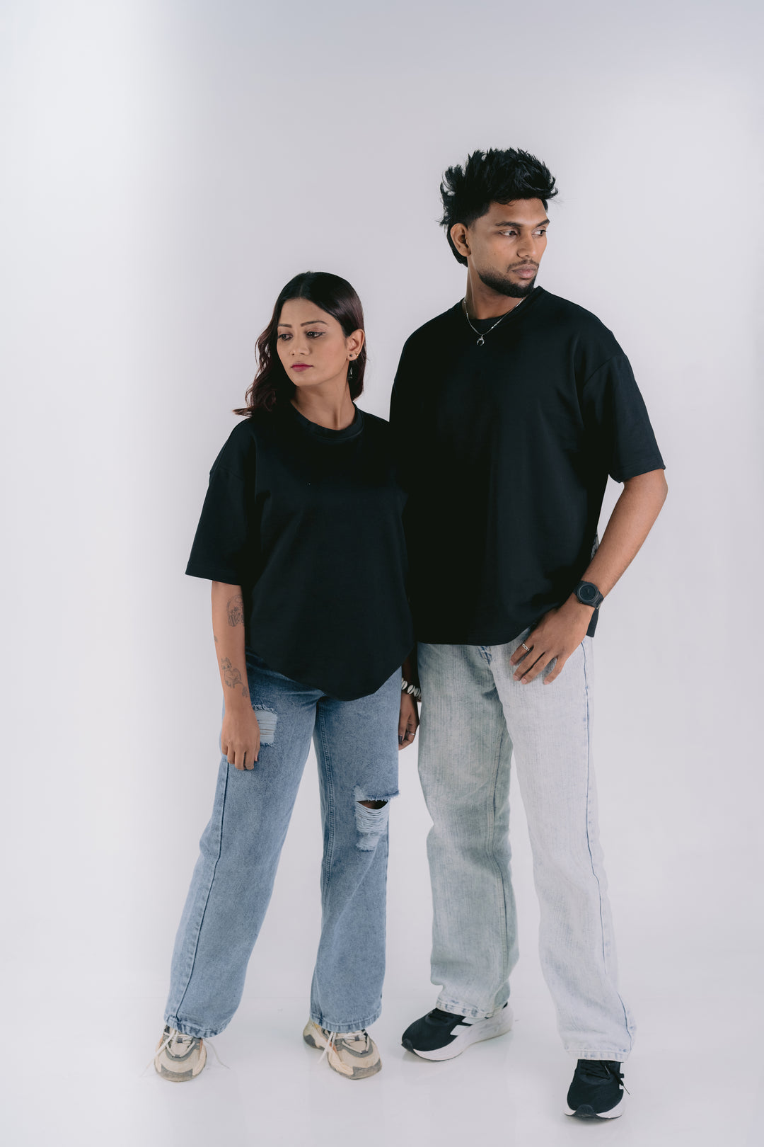 A man and a woman modeling oversized black t-shirts and light-wash jeans against a plain light gray studio background.