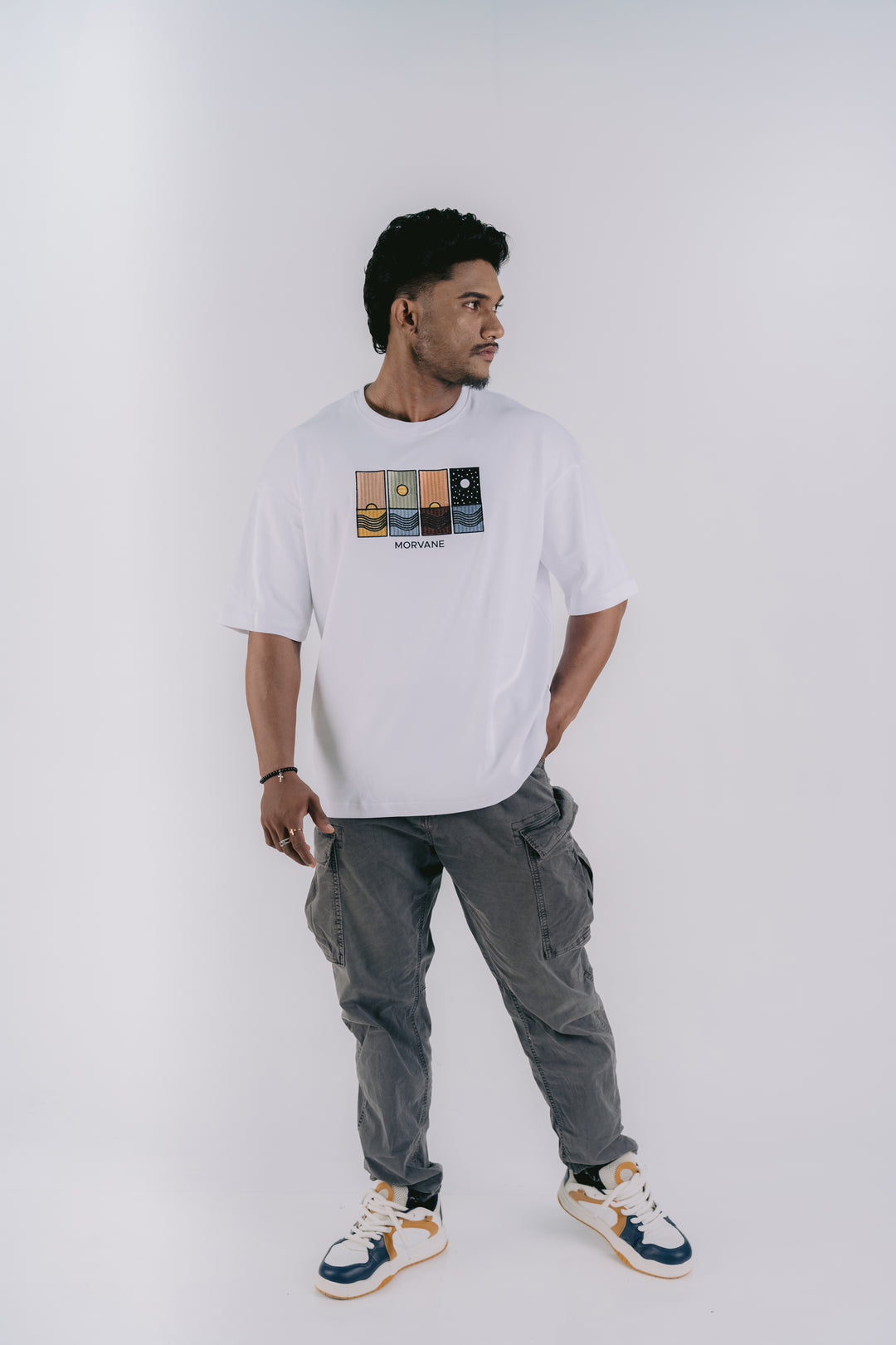 A man modeling a white crewneck t-shirt with a graphic print on the chest, paired with gray cargo pants and white, blue, and orange sneakers, set against a plain light gray studio background.
