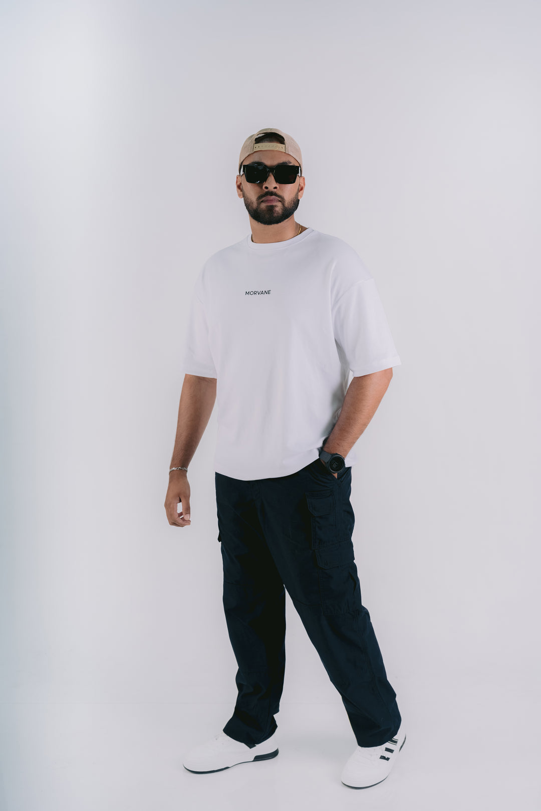 A man modeling a white crewneck t-shirt with a small black logo on the left chest, paired with dark cargo pants, white sneakers, and a beige baseball cap, against a plain light gray background.