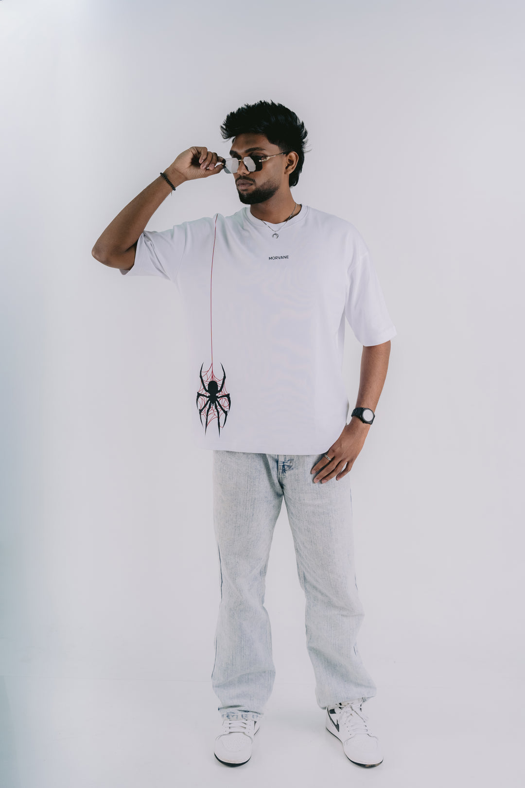 A man wearing a white t-shirt with a black spider graphic and a matching spider-shaped bag, styled with light wash jeans and white sneakers against a plain white background.