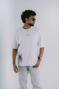A man wearing a white short-sleeve t-shirt with a black spider graphic on the lower left and small black text on the chest, paired with light-wash jeans, photographed against a light gray studio background.