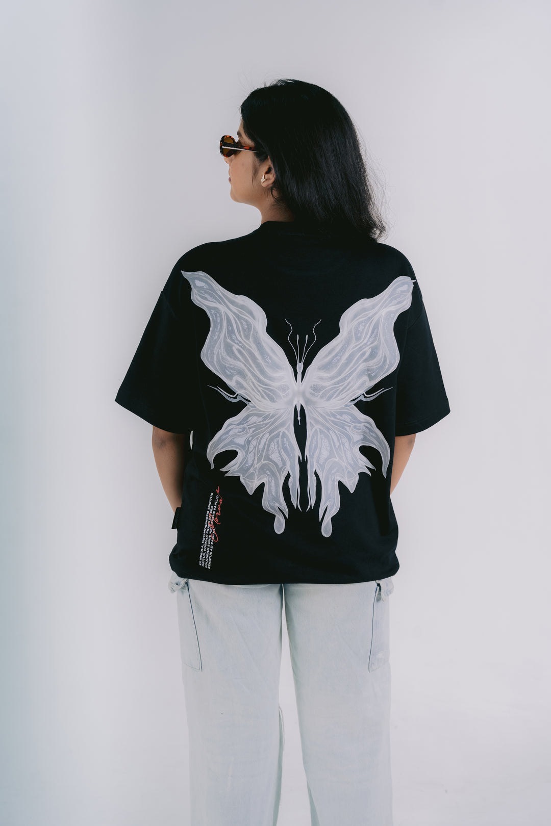 A woman viewed from behind wearing a black oversized T-shirt with a large white butterfly graphic on the back, paired with light gray pants, against a plain light gray studio background.