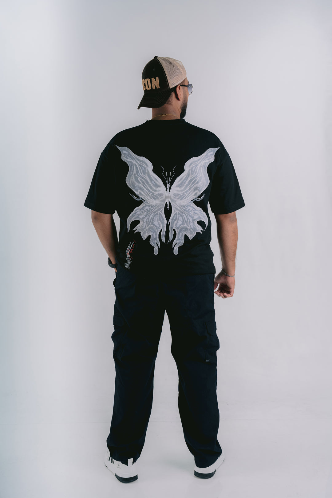 A man viewed from behind wearing a black t-shirt with a large white butterfly graphic on the back, black cargo pants, and a black and beige trucker hat.