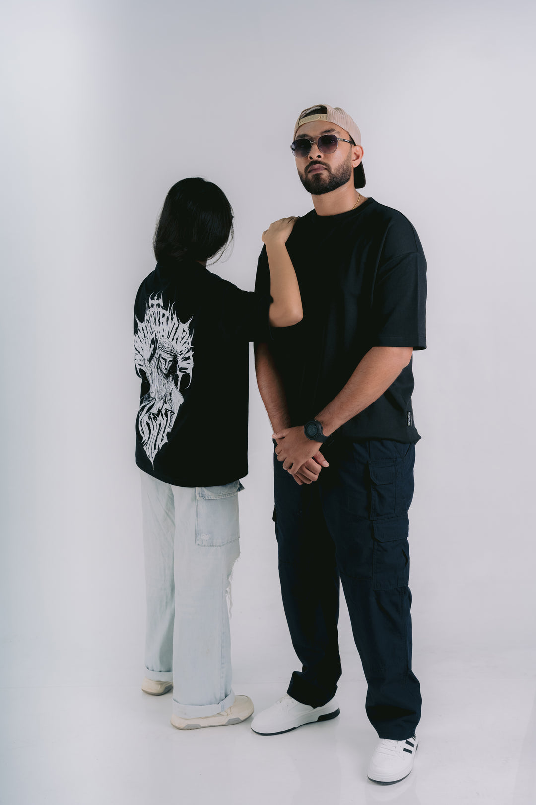 Two models in a studio setting, one showing the back of a black graphic t-shirt with a white skull design, and the other wearing a black crewneck t-shirt, dark cargo pants, and a beige cap.