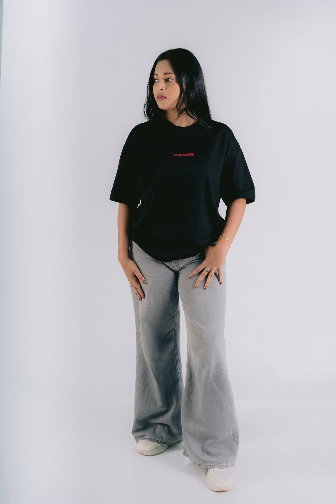 A woman modeling a black oversized t-shirt with a small red text graphic on the chest, paired with wide-leg light gray sweatpants and white sneakers, standing against a plain white studio background.